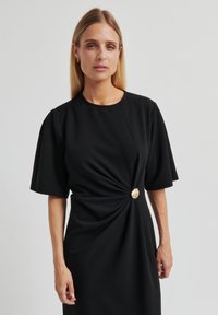 Robe noire avec manches courtes bouffantes, détail torsadé à l'avant et accent circulaire doré à la taille. Confectionnée en tissu lisse.