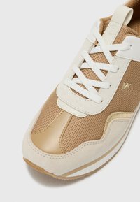 Beige Sneaker mit weißen Akzenten, aus Netz- und Ledermaterialien, mit einer abgerundeten Zehenpartie, kontrastierenden Texturen und einem goldenen Logo-Detail.