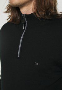 Schwarzer Strickpullover mit hohem Kragen und einem Viertelreißverschluss vorne, mit grauer Stickerei und einem kleinen weißen "ck"-Logo auf der Brust.