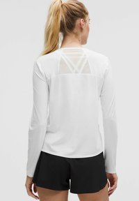 Bijela majica dugih rukava s prozirnom mrežastom stražnjom pločom koja ima geometrijski uzorak, uparen sa crnim šortsevima. Glatka tekstura i sportski kroj.