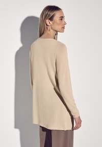 Maglietta beige a maniche lunghe realizzata in tessuto testurizzato, con scollatura rotonda e una vestibilità morbida, mostrata di profilo con accessori minimi.