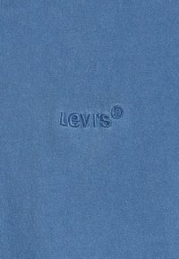 Modrá bavlněná tkanina s hladkou strukturou má embosované logo "Levi's". Materiál je lehký a flexibilní, ideální pro volnočasové nošení.