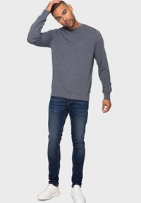 Grauer Sweatshirt mit rundem Ausschnitt und gerippten Bündchen, kombiniert mit dunkelblauen Skinny Jeans und weißen Turnschuhen. Das Modell posiert mit einer Hand am Kopf.