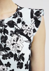 Blusa senza maniche di colore azzurro chiaro con pieghe e stampa floreale bianca e nera indossata da una persona.