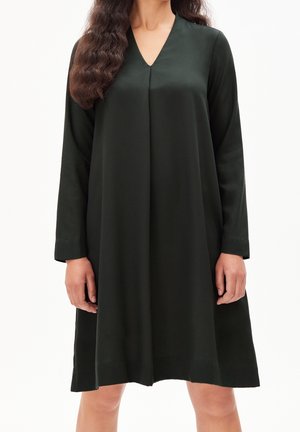 Frau trägt ein locker sitzendes, langärmliges, knielanges schwarzes Kleid mit V-Ausschnitt.