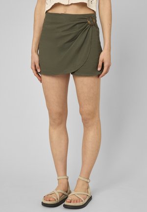 Femme portant un short style portefeuille vert olive avec un détail anneau en écaille et des sandales beige à bride cheville sur fond gris.