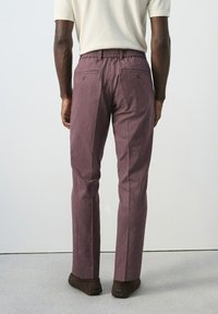 Hombre vistiendo pantalones marrón granate con rayas finas, camisa blanca de manga corta y zapatos marrón oscuro, de pie contra un fondo liso.