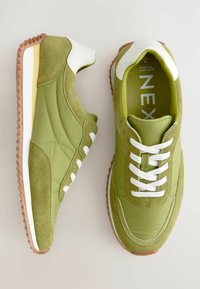 Grønne sneakers med en kombination af glatte og ruskindsteksturer, hvide snørebånd og en gummisål med et zigzag-mønster. Distinktive design detaljer er synlige.