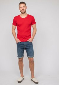 T-shirt rouge à col en V à manches courtes associé à un short en denim roulé ; le mannequin porte des chaussures à enfiler crèmes, mettant en valeur un look d'été décontracté.