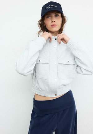 Mlada ženska, ki si prilagaja svetlo sivi krajšani jakni s pritisnimi gumbi, oblečena v mornarsko modro krilo in mornarsko modro kapo z belim napisom "San Francisco EST 1994."