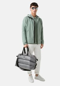 Lightweight mint green jacket with a hood, paired with a darker green shirt, white pants, and gray puffer-style duffel bag with black accents.