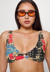 Top de biquíni multicolorido com padrão floral em vermelho, azul e preto, com um detalhe central em anel dourado e alças largas.