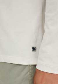 Weißes Langarmhemd mit Rundhalsausschnitt, versehen mit einem kleinen blauen und grauen Logopatch auf der linken Unterseite und einer weichen Textur.