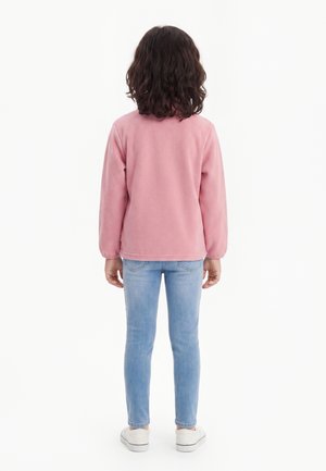 Child with curly dark hair stands facing away wearing a pink long-sleeve top, light blue jeans, and white sneakers.