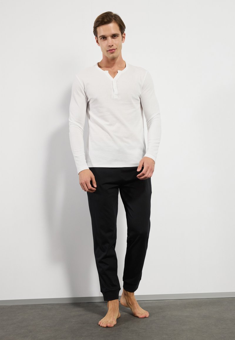 Hvid langærmet henley skjorte med tre-knaps placket, parret med sorte bukser med elastisk talje, præsenteret på en model uden sko.