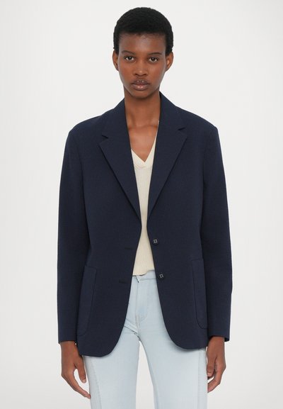 Navy pinstriped blazer with notch lapels, two front pockets, and button closures, worn over a light-colored top and paired with jeans.
