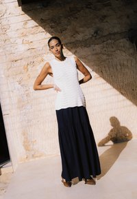 Femme debout, mains sur les hanches, contre un mur beige rugueux éclairé par le soleil, portant un haut en maille blanc sans manches, une longue jupe noire et des chaussures marron.