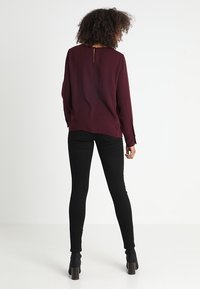 Blouse bordeaux à manches longues avec une ouverture en forme de trou à l'arrière, associée à un jean skinny noir et des bottines à talons, vue de dos.