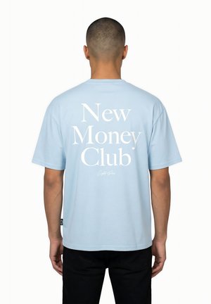 Uomo che indossa una maglietta azzurro chiaro con una grande scritta bianca "New Money Club" sulla schiena, abbinata a pantaloni neri, di spalle.