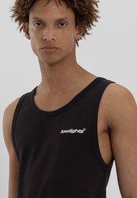 Ung person med krøllet hår iført en sort tanktop med et lille hvidt "lowlights" logo på brystet, som står fremadrettet mod en ensfarvet baggrund.