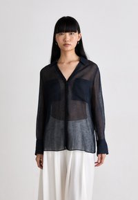 Mulher vestindo uma blusa preta transparente de manga longa com dois bolsos frontais, combinada com calças brancas largas, contra um fundo claro e simples.