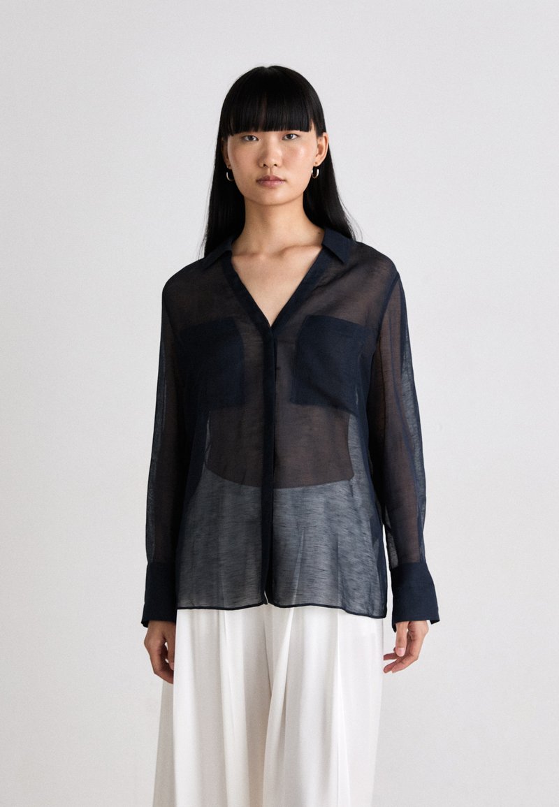 Mulher vestindo uma blusa preta transparente de manga longa com dois bolsos frontais, combinada com calças brancas largas, contra um fundo claro e simples.