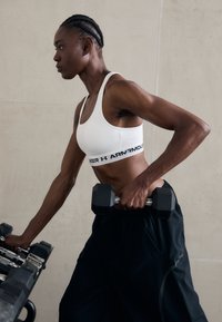 Kvinna i vit sportbehå och svarta byxor som håller en hantel och väljer vikter från en stång i ett gym.