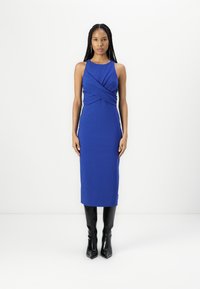 Rochie fără mâneci, de culoare albastră, cu un detaliu răsucit la talie, siluetă ajustată, până la genunchi, purtată împreună cu cizme negre cu toc înalt.