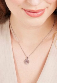 Zilveren hangerketting met een ronde charme met een boomontwerp, bevestigd aan een delicate ketting, gedragen boven een lichtbeige top.