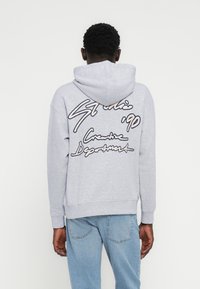 Γκρι φούτερ με κουκούλα και κορδόνι, με μεγάλο, στυλιζαρισμένο κείμενο στην πλάτη σε σκούρο μπλε και ανοιχτό μπεζ χρώμα. Ριμπ μανσέτες και τελείωμα.