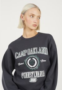 Sweatshirt gris foncé avec un col rond affichant le texte blanc et vert "CAMP OAKLAND" et des graphiques. Texture douce, coupe ample et poignets côtelés.