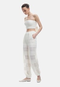 Weißes Zweiteiler-Outfit mit einem gerafften, trägerlosen Oberteil und transparenter, gemusterter weit geschnittener Hose. Der Stoff ist leicht und strukturiert.