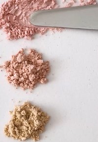 Pigmenty w proszku w jasnoróżowym, średniopróżowym i beżowym kolorze, o gładkiej teksturze, ułożone na białej powierzchni obok szpatułki.