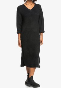 Langes schwarzes Strickkleid mit V-Ausschnitt und dreiviertel Ärmeln, mit Rippenstruktur und Seitenschlitzen. Kombiniert mit schwarzen Stiefeletten.