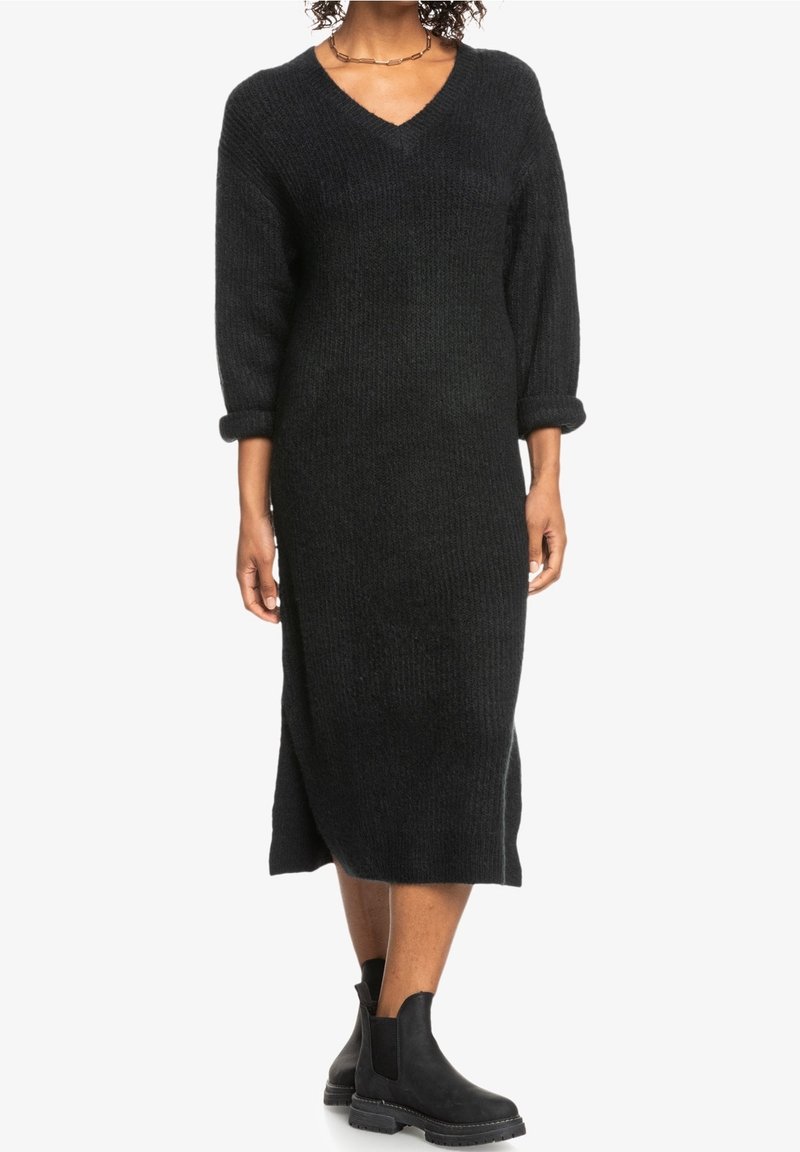 Langes schwarzes Strickkleid mit V-Ausschnitt und dreiviertel Ärmeln, mit Rippenstruktur und Seitenschlitzen. Kombiniert mit schwarzen Stiefeletten.