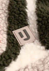 Texturovaná tkanina s zelenými, růžovými a bílými pruhy a obdélníkovou šedou etiketou s černým logem "J". Měkký a chlupatý vzhled.