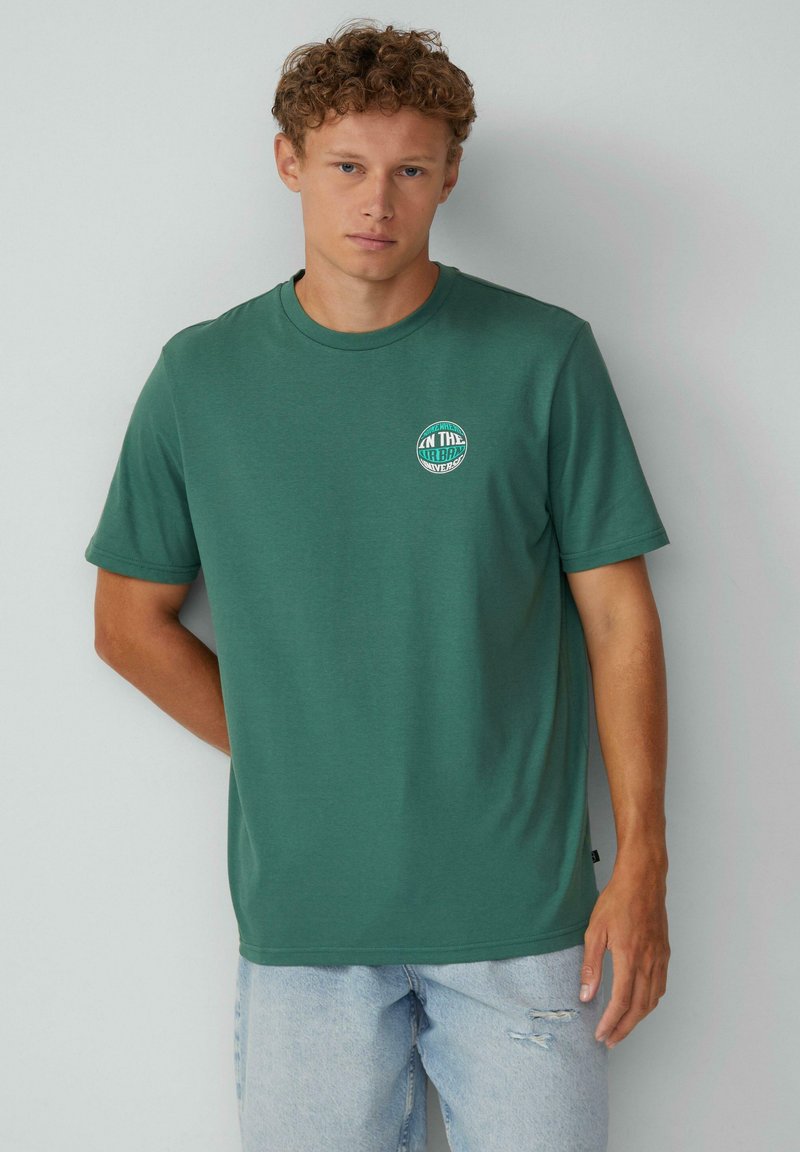 Groene korte mouwen t-shirt van zachte stof, met een ronde hals en een kleine ronde afbeelding op de linkerborst. Draagt met lichtblauwe versleten jeans.