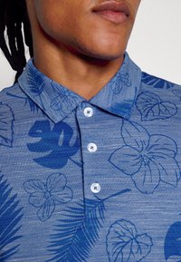 Lähedalt vaade inimesest, kellel on seljas sinine nööpidega särk, tumedate siniste troopiliste lillemuster ja krae.