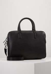 Porte-documents en cuir noir avec doubles poignées, bandoulière détachable et détails en fermeture éclair argentée, posé sur une surface blanche avec un fond uni.