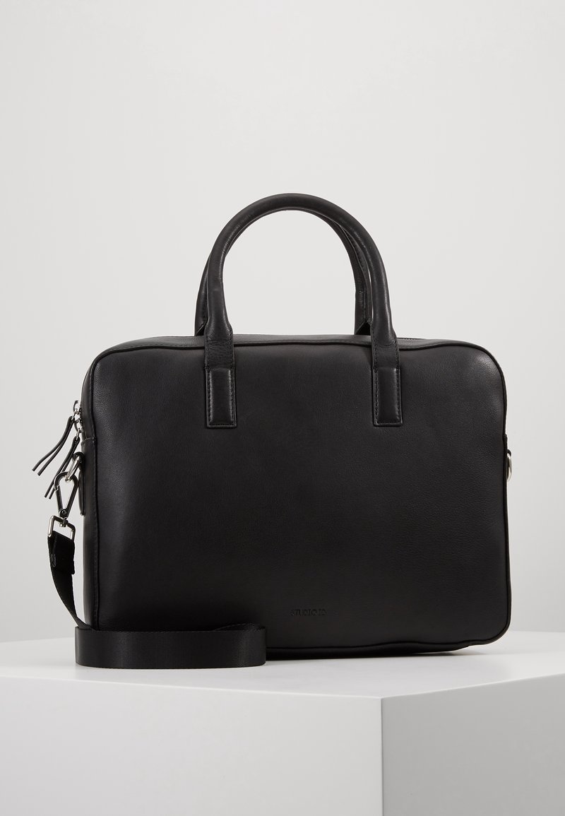 Porte-documents en cuir noir avec doubles poignées, bandoulière détachable et détails en fermeture éclair argentée, posé sur une surface blanche avec un fond uni.