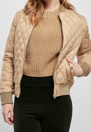 Femme portant une veste bomber matelassée beige sur un pull en maille beige et un pantalon noir taille haute, une main dans la poche de la veste.
