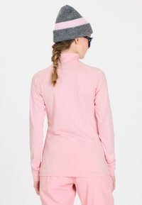 Helles rosa Langarmoberteil mit hohem Kragen und Daumenlöchern. Kombiniert mit einer grauen Mütze mit rosa Streifen. Glatte Textur, sportliche Passform.