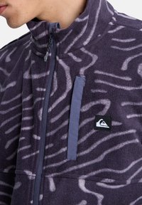Fleece jacket in dark purple with white wavy patterns, featuring a half-zip, high collar, and a zippered chest pocket with logo.