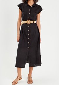 Femme portant une robe midi noire boutonnée sur le devant, à manches courtes, avec deux poches poitrine, une ceinture beige et des sandales plates marron.