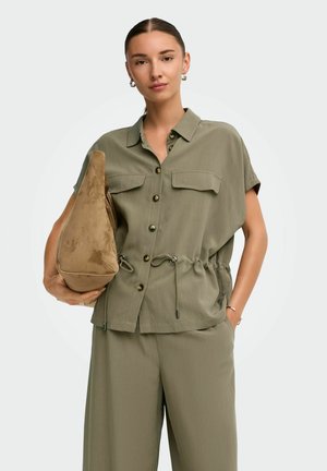 Joven con el pelo echado hacia atrás sostiene un gran bolso de ante color beige, lleva una camisa verde oliva abotonada y pantalones anchos a juego.