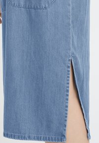 Jupe en denim bleu clair avec une fente latérale dévoilant une partie de la jambe, mettant en valeur la texture du denim et les détails des coutures.