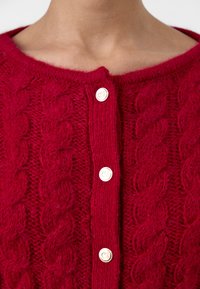 Cardigan rouge en tricot avec motif en cable, col rond et trois grands boutons crème sur le devant. Texture douce et apparence chaleureuse.