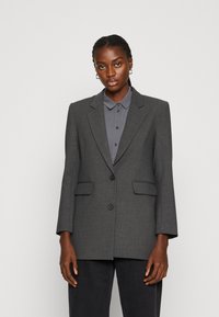 Grijze op maat gemaakte blazer met notch-klepels, enkele knoopsluiting, twee voorzakken en gestructureerde schouders, gedragen over een grijs overhemd met kraag.