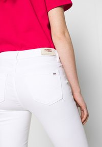 Jeans de denim blanco con un diseño ajustado, que presentan bolsillos traseros y una etiqueta de Tommy Jeans en la cinturilla. Acompañados de una blusa de color rosa brillante.