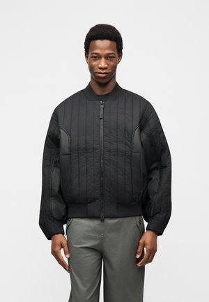 Άνδρας που φορά μαύρο μπουφάν τύπου bomber με φερμουάρ και ραμμένες επενδύσεις, στέκεται μπροστά σε απλό ανοιχτό φόντο, με τα χέρια χαλαρά δίπλα στο σώμα.